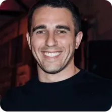 Smiling man with short dark hair wearing a black shirt against a dark indoor background.